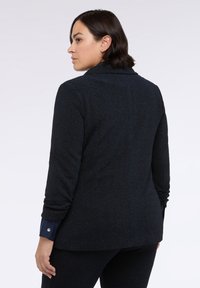 Černý texturovaný blejzr s dlouhými rukávy, klopovým límcem a lesklým detailem na manžetách, viděný ze zadního úhlu.