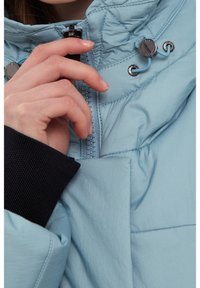 Giacca isolante azzurro chiaro con chiusura zip, hardware argentato e dettagli elastici. Tessuto nero testurizzato visibile ai polsini.