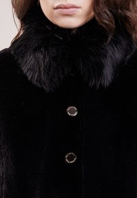 Sort pel coats med en blød krave, tekstureret stof og runde metalliske knapper med indgraveret branding i midten.