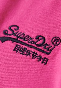 Logo "SuperDry" nero ricamato e caratteri giapponesi su tessuto rosa acceso, primo piano del dettaglio dell'abbigliamento.