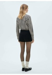 Zwarte shorts gecombineerd met een gestreepte langemouwen top. Het model draagt doorzichtige panty's, crèmekleurige kniesokken en zwarte schoenen. Effen achtergrond.