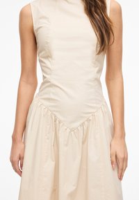 Femme portant une robe crème sans manches avec un corsage ajusté et une jupe froncée, debout sur un fond blanc uni.