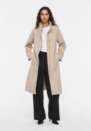 Une femme porte un long manteau beige sur un haut blanc et un pantalon noir, devant un fond de studio sobre.