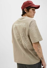 Beige Baumwoll-T-Shirt mit kurzen Ärmeln, das auf der Rückseite ein weißes Blattdesign und Markentext zeigt. Kombiniert mit einer kastanienbraunen Mütze.