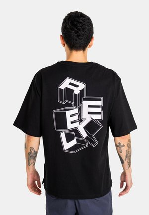 Mann mit kurzen schwarzen Haaren und Tätowierungen an den Armen trägt ein schwarzes oversized T-Shirt mit weißen 3D-Blockbuchstaben, die auf dem Rücken „RECK“ schreiben.