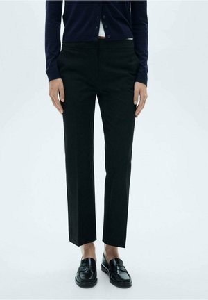 Person iført korte sorte lige bukser, sorte loafers og en navy cardigan med hvid top synlig under.