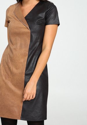 Frau trägt ein kurzärmeliges, knielanges Kleid mit einem vertikalen Schnitt aus beigem Wildleder und schwarzem Kunstleder.