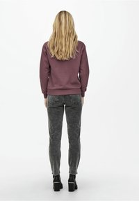 Bordeauxroter Sweatshirt mit gerippten Bündchen und Saum, kombiniert mit hochgeschnittenen, ausgewaschenen schwarzen Jeans. Modell trägt schwarze Absatzschuhe, von hinten betrachtet.