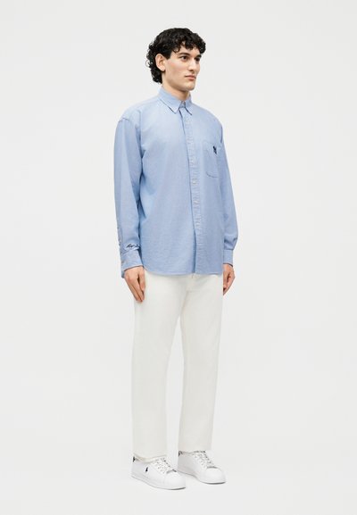 Άνδρας που φοράει ανοιχτό μπλε πουκάμισο με κουμπιά, λευκό παντελόνι και λευκά αθλητικά παπούτσια, στέκεται μπροστά σε απλή λευκή φόντο.