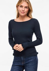 Femme portant un pull ajusté côtelé bleu marine avec des poignets et un ourlet à volants, associé à un jean bleu foncé, debout devant un fond uni.