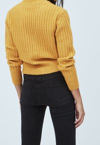 Kvinna i en senapsgul ribbad tröja och tighta svarta jeans, sedd bakifrån mot en neutral ljus bakgrund.