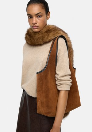 Brązowa torba typu tote ze skóry zamszowej z czarną skórzaną lamówką, noszona na beżowym swetrze z brązowym futrzanym kołnierzem, na neutralnym tle.
