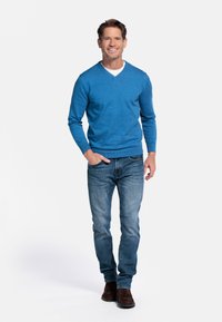 Blauwe v-hals trui met een gestructureerd patroon, gecombineerd met vervaagde spijkerbroeken en donkerbruine laarzen. Model staat tegen een witte achtergrond.