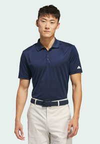 Marineblauw poloshirt met een kraagontwerp en drie knopen, gecombineerd met witte broek en een gestructureerde blauwe riem. Inclusief het Adidas-logo op de mouw.