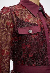 Μπλούζα μπορντό από δαντέλα με λουλουδάτο μοτίβο, σταθερό γιακά και πατιλέτα τσέπης στερεωμένη με περίτεχνα ασημένια κουμπιά, φορεμένη από άτομο.