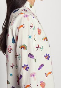 Camisa de tela ligera con fondo crema y patrones abstractos coloridos, que incluyen animales, plantas y formas geométricas.