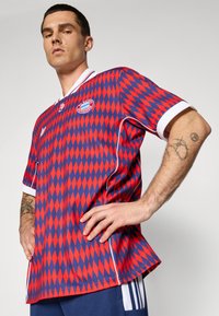 Czerwona i niebieska koszulka piłkarska w diamentowy wzór z białym kołnierzykiem i rękawami, z logo i paskowanymi akcentami, wykonana z tkaniny.
