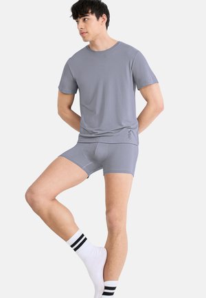 Man wearing gray short-sleeve shirt and matching boxer briefs, standing on one leg with hands behind back against plain background.