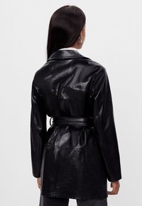 Czarny skórzany płaszcz przeciwdeszczowy z paskiem w talii, długimi rękawami i gładką fakturą. Posiada dopasowany krój oraz subtelny połysk.