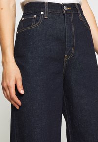 Dark blue denim jeans with a high waist, featuring copper stitching, five-pocket design, and metal buttons. The texture is slightly grainy.
