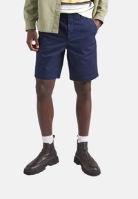 Pantalones cortos de algodón color navy con bolsillos laterales, combinados con botas de cuero marrón oscuro hasta el tobillo y calcetines de canalé blancos, estilizados con una chaqueta verde claro.