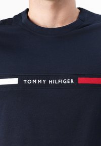 Tengerészkék pamut póló kerek nyakkivágással, fehér és piros vízszintes csíkokkal, valamint a "TOMMY HILFIGER" felirattal fehér szöveggel.
