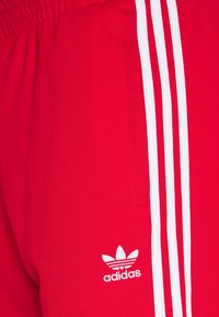 Červené sportovní kalhoty vyrobené z měkkého, elastického materiálu s bílým trojitým pruhem a malým bílým logem na levé straně.