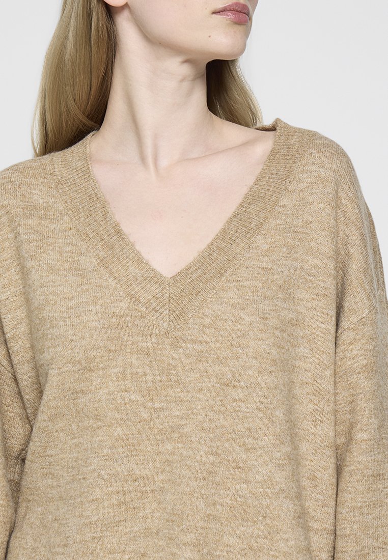 Suéter de punto beige con diseño de escote en V, ajuste holgado y tejido texturizado. Presenta puños y dobladillo acanalados, lo que enfatiza su apariencia suave.