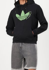 Svart hoodie med en pixelerad grön adidas trefoil-logga. Tyget har en mjuk textur, med en känguruficka och ribbade muddar.