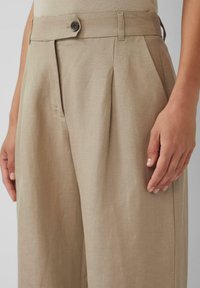 Khaki-Hosen mit Falten, einem strukturierten Stoff und einem einzelnen dunklen Knopf an der Taille. Eine Hand, die an der Seite ruht, fügt Detail hinzu.