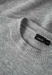Pull gris tricoté avec un col côtelé, présentant une texture lisse et des coutures visibles. L'étiquette de la marque indique "LMTD", taille 13-14.
