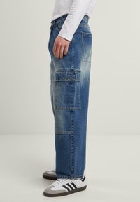 Blue denim cargo pants with a loose fit, multiple pockets, and a faded wash, paired with white athletic shoes featuring black stripes.
