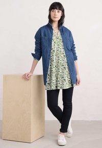 Eine Jeanshemdjacke über einer floral gemusterten Tunika in grün-weißen Farben, kombiniert mit schwarzen Skinny-Jeans und weißen Sneakers vor einem neutralen Hintergrund.