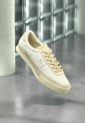 Sneaker casual bianca e beige con lacci e suola testurizzata, che fluttua sopra un pavimento grigio riflettente vicino a una parete di vetrocemento.