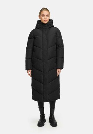 Longue doudoune noire avec capuche, design matelassé avec motifs en chevrons, coupe droite et poches latérales, portée sur un pantalon noir et des bottes.