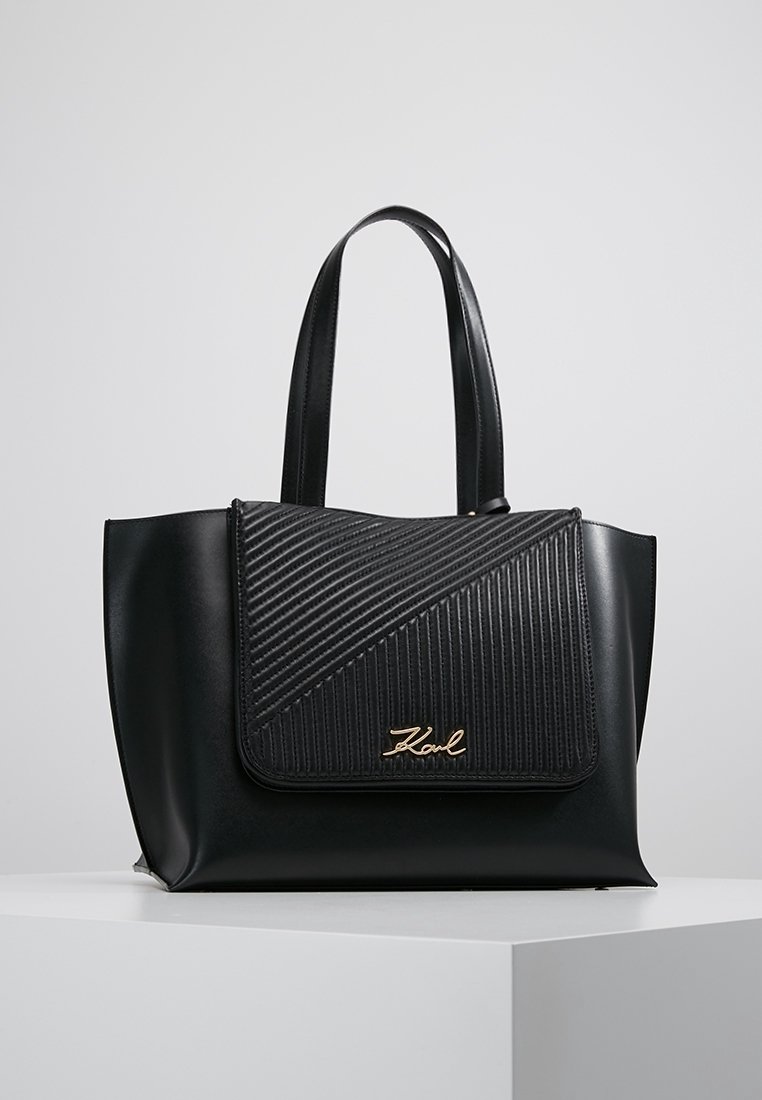 Sac fourre-tout noir en simili cuir lisse avec un rabat texturé à côtes diagonales. Présente des poignées à double portage et un accent de logo doré.