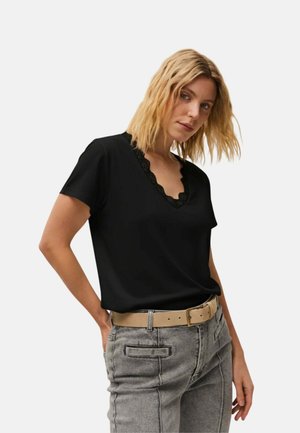 Mujer rubia que lleva una camiseta negra de manga corta con escote de encaje metida en unos jeans grises de tiro alto con un cinturón beige.