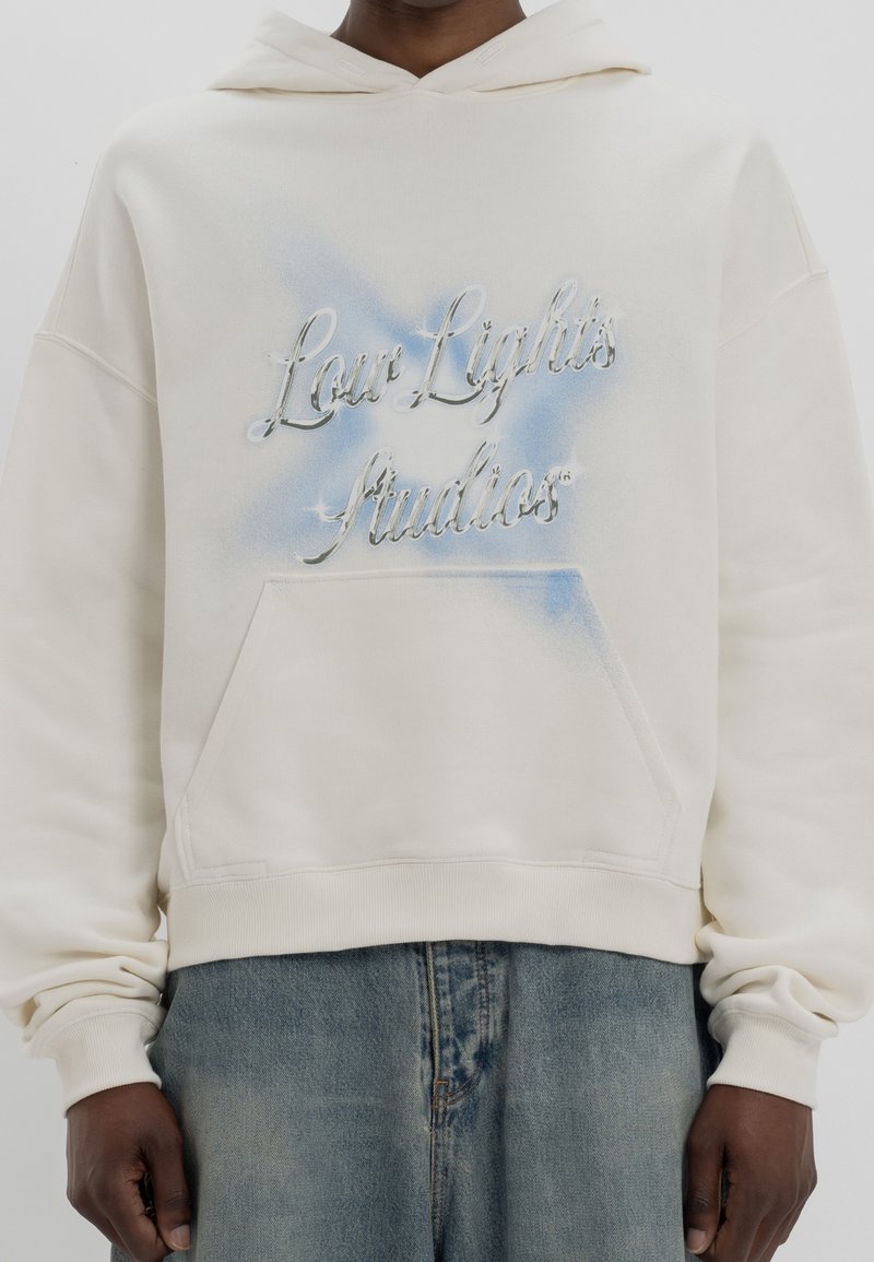 Sweat à capuche couleur crème avec une poche avant, affichant un texte dégradé bleu "Low Lights Studios" en police métallique. Texture de tissu doux.