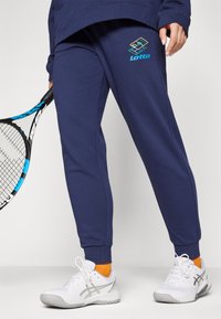 Mørkeblå joggebukser i mykt materiale med brodert Lotto-logo. Kombinert med hvite joggesko og oransje sokker.