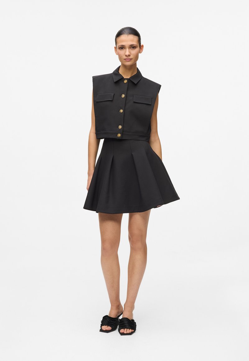 Top negru fără mâneci, scurt, cu nasturi aurii și două buzunare, asortat cu o fustă neagră evazată. Ținuta este realizată dintr-un material neted.