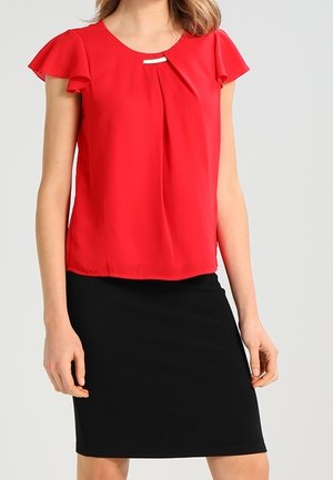 Haut rouge à manches courtes avec accents en volants, devant plissé, et détail en métal argenté au niveau de l'encolure, associé à une jupe noire moulante.