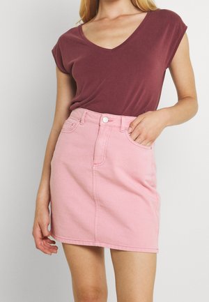 Mujer que lleva una camiseta burdeos con cuello en V metida en una minifalda vaquera rosa de talle alto, con la mano en el bolsillo.