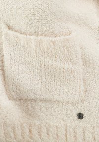 Cremefarbener weicher Fleece-Stoff mit einer quadratischen Tasche und metallicfarbenem Knopfdetail, das eine strukturierte Oberfläche und unregelmäßige Strickmuster aufweist.