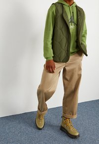 Gilet trapuntato verde sopra una felpa verde, abbinato a pantaloni beige e stivali verde oliva con suole nere e lacci arancioni.