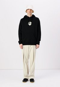 Μαύρη κουκούλα με μπροστινή τσέπη και λογότυπο, μπεζ καπέλο τύπου bucket, ανοιχτόχρωμο παντελόνι και μαύρα αθλητικά παπούτσια. Απλός σχεδιασμός, άνετη εφαρμογή.