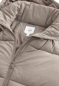 Puffjacke aus leichtem Taupe-Stoff, mit hohem Kragen, Reißverschluss und einer glatten, leicht knitterigen Textur. Innenetikett sichtbar.