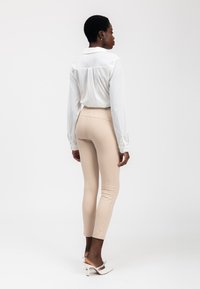 Witte lange mouwen shirt, aansluitende beige leggings en witte hoge hakken sandaaltjes. Het shirt heeft een kraag; de leggings hebben een gladde textuur. Achteraanzicht.