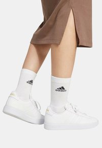 Sneakers bianchi a basso profilo con tre strisce laterali, dotati di una tomaia in pelle liscia e dettagli dorati. Abbinati a calzini bianchi a coste.