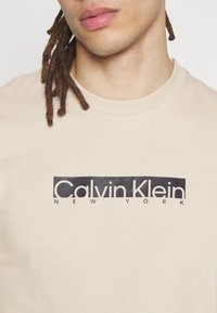Mladý člověk s dredy, oblečený v krémovém tričku s kulatým výstřihem a černým logem "Calvin Klein New York" na hrudi.