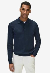 Marinblå långärmad pikétröja med ribbat mönster, krage, knappstängning och ribbade ärmslut. Bärs med ljusa byxor.